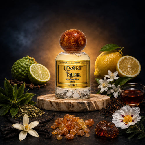 A bottle of Levant Musk eau de parfum ABEL centered on a wood slice, surrounded by lemons, a lime, flowers, vanilla beans, amber crystals, and a small glass of dark liquid on a dark smoky background.