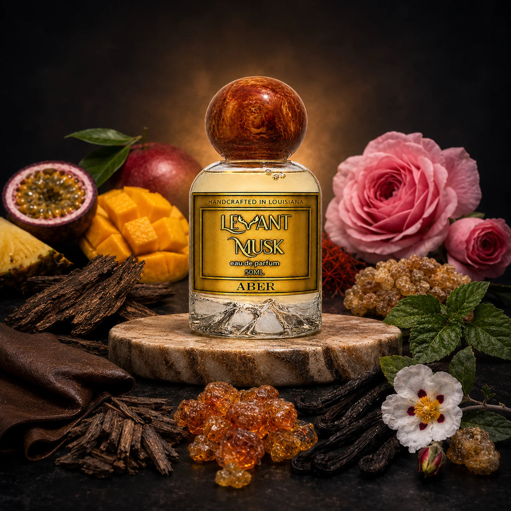 A bottle of Levant Musk ABER eau de parfum stands on a round marble slice, surrounded by botanical and flavor-inspired props including pink roses, a passion fruit, mango pieces, vanilla beans, amber crystals, and dark brown fabric against a dark background.