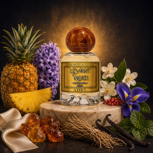 A perfume bottle labeled Adel, by Levant Musk styled on a marble pedestal with pineapple, purple flowers, white blossoms, vanilla beans, mint, amber crystals, and a cream ribbon against a dark smoky background.