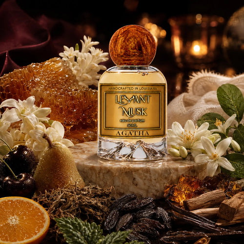 A bottle of Agatha by Levant Musk eau de parfum is centered on a marble slab, surrounded by rich, warm fragrance notes including white flowers, a pear, vanilla pods, amber, and citrus, with a softly blurred candlelit background.