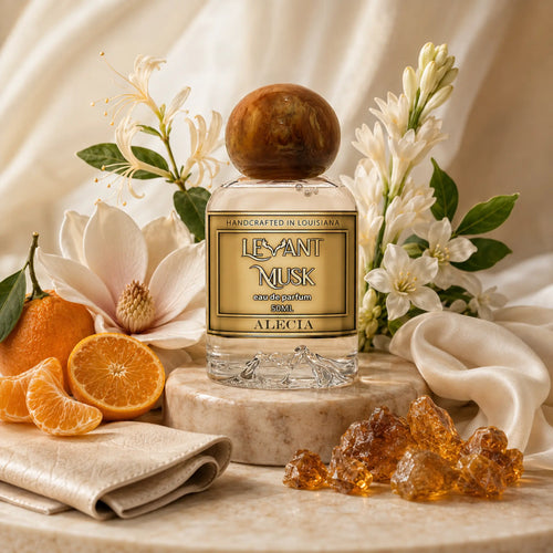 An eua de parfum 50ml bottle labeled ALECIA, by Levant Musk styled on a marble pedestal with white flowers, oranges, amber resin, and cream fabric in a warm, elegant composition.