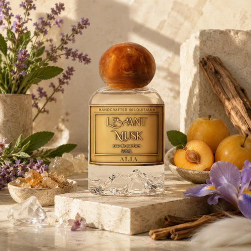 A clear glass perfume bottle labeled ALIA by Levant Musk with a round amber-brown  cap, styled on a stone pedestal with apricots, purple flowers, crystals, and dried botanicals in a warm rustic setting.