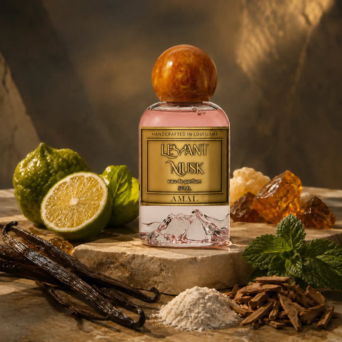 A bottle of Amal eau de parfum by Levant Musk stands on a stone slab surrounded by its natural ingredients, including a halved lime, vanilla pods, mint leaves, and resin pieces, under warm, dramatic lighting.