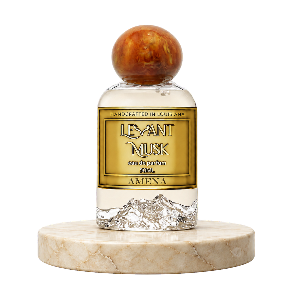 A 50ml bottle of Amena by Levant Musk eau de parfum, featuring a clear glass body with a gold label, a textured mountain design at the base, and a round, marbled amber-brown cap, displayed on a round marble pedestal against a white background.