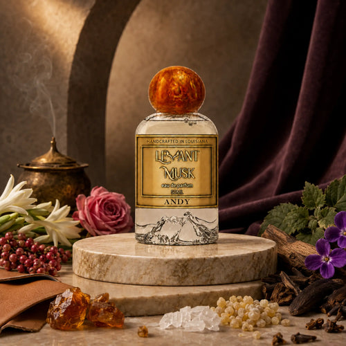 A perfume bottle labeled Andy eau de parfum by Levant Musk stands on a round stone pedestal. The bottle has a gold label, clear glass, and a round amber-colored top
