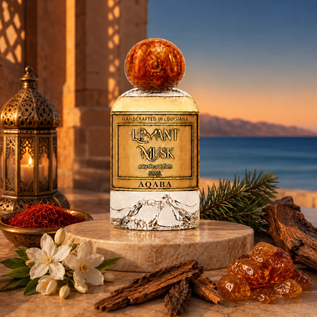 A perfume bottle labeled Aqaba, by Levant Musk styled on a stone pedestal with saffron, jasmine flowers, cinnamon sticks, amber crystals, and a lantern against a sunset seascape background.