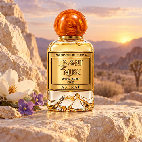ASHRAF by Levant Musk perfume displayed with magnolia and violet flowers on pale desert rocks at sunset.