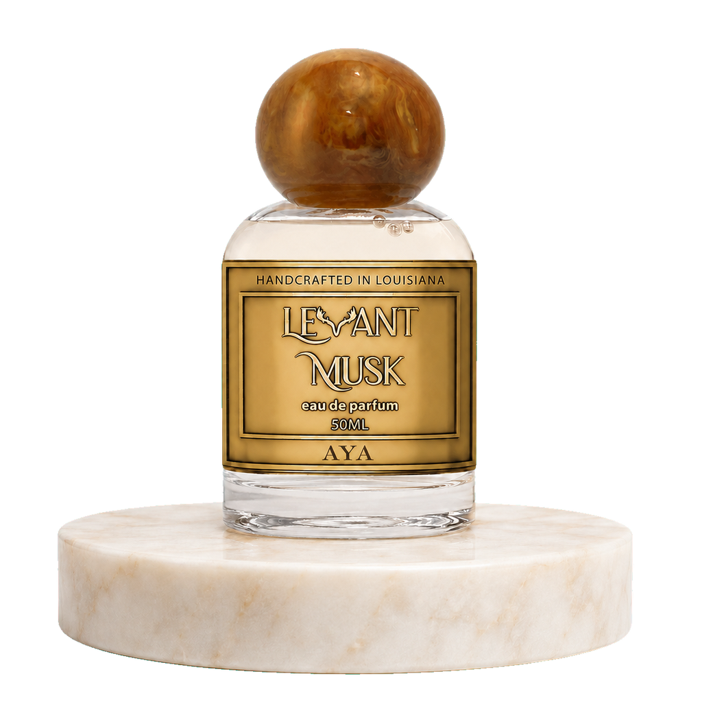 A 50ml bottle of AYA by Levant Musk Eau de Parfum, featuring a clear glass body, a gold label with black lettering, and a polished brown wooden stopper, displayed on a white marble pedestal against a white background.