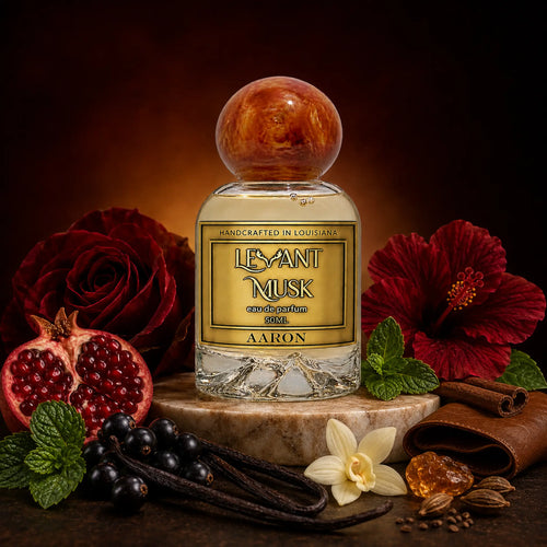 A bottle of eau de parfum Aaron by Levant Musk, styled with red roses, a pomegranate, vanilla beans, cinnamon, and a white orchid on a marble pedestal against a dark background.