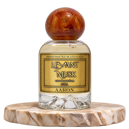 A bottle of Levant Musk Eau de Parfum labeled Aaron, featuring a clear glass bottle with a round amber-brown wooden cap and a gold label, displayed on a round marble coaster against a white background.
