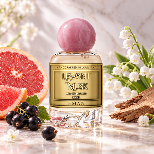 A bottle of Eman Levant Musk eau de parfum centered on a marble surface, surrounded by fragrance-inspired props including a sliced grapefruit, blackcurrants, lily of the valley flowers, and wood bark. The bottle has a pink spherical cap and a gold label with black text.