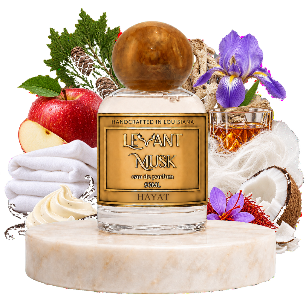 A bottle of Hayat by Levant Musk handcrafted fragrance  in a clear glass bottle with a round amber cap, displayed on a marble pedestal with scent notes including an apple, folded white towels, purple irises, coconut, and a glass of amber liquid.