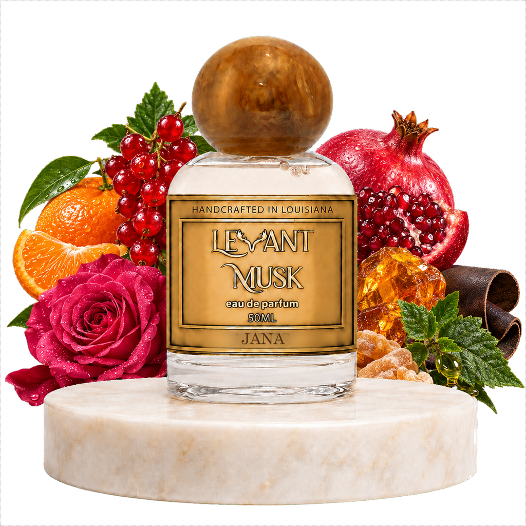 A clear glass perfume bottle with a round AMBER cap and a gold label reading 'LEVANT MUSK' and 'JANA', displayed on a white marble pedestal with fruit and flowers around it, including a pomegranate, orange slices, red berries, a pink rose, amber honey, cinnamon, and mint, on a white background.