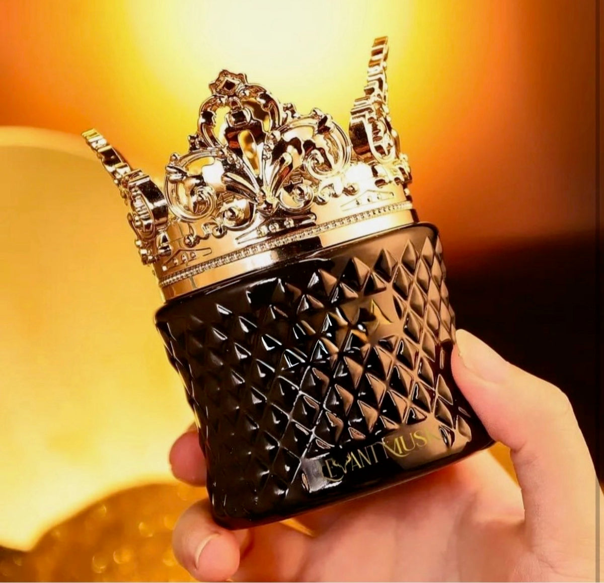 A hand holding a black textured cosmetic bottle with a metallic gold crown cap, featuring the brand name 'Levant Musk' on the front, set against a warm glowing background.