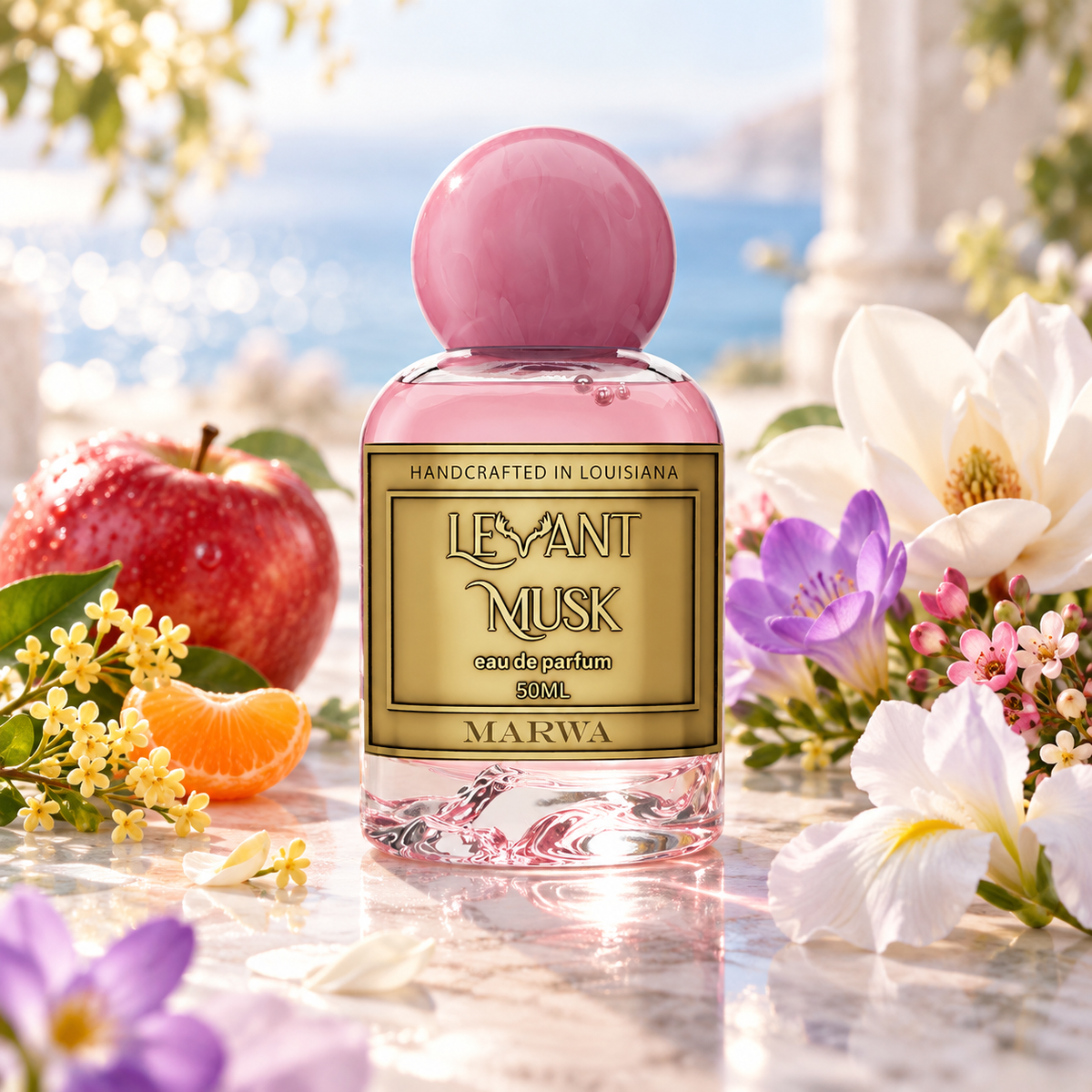 A 50ml bottle of Marwa Levant Musk Eau de Parfum is centered on a marble surface, surrounded by fresh flowers, a red apple, and an orange slice, with a blurred outdoor sea and garden background.