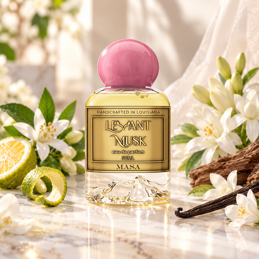 A bottle of Masa by Levant Musk Eau de Parfum , featuring a pink spherical cap and a gold label, styled on a marble surface with white flowers, a lime, and a vanilla bean.