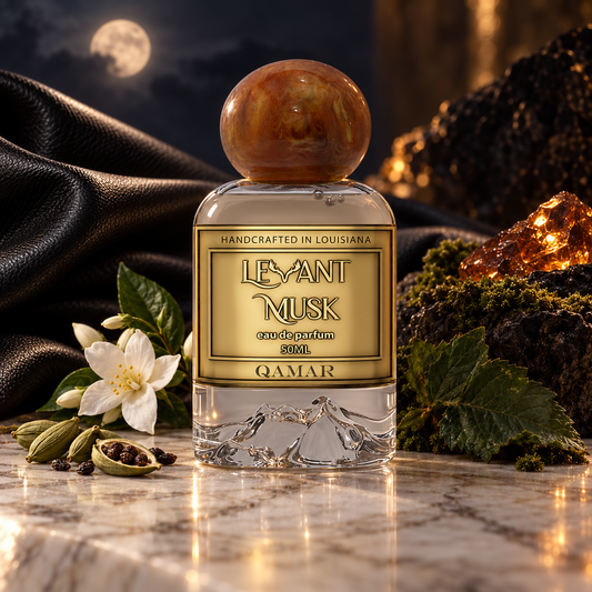 A clear glass perfume bottle labeled  QAMAR, by 'LEVANT MUSK' with a round amber-brown cap, set on a marble surface with a white flower, green leaves, spices, black fabric, and glowing amber stones in a moody styled scene.