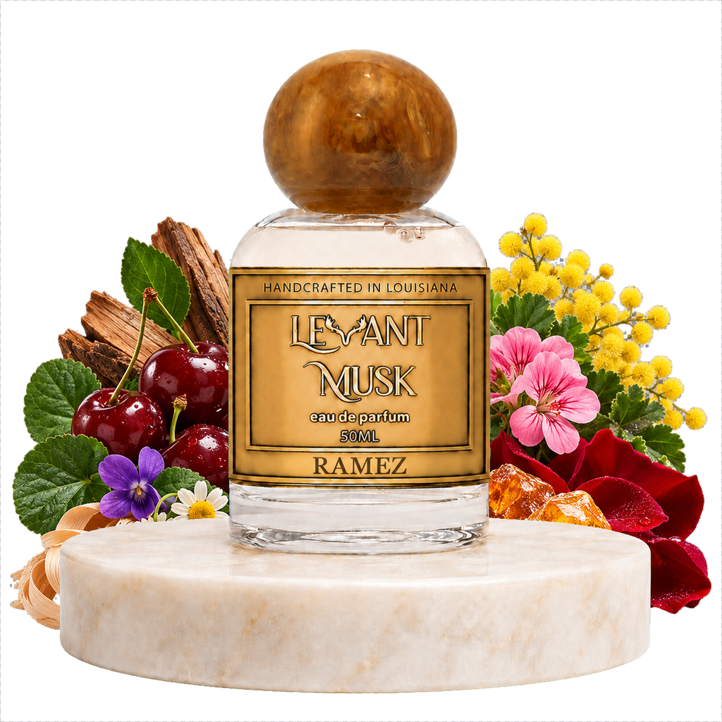 A clear glass perfume bottle with a round polished amber cap and a gold label reading 'LEVANT MUSK' EAU DE PARFUM NAMED 'RAMEZ,' displayed on a white marble pedestal with cherries, flowers, and wood pieces around it on a white background.