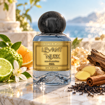A bottle of Ruston Levant Musk Eau de Parfum stands on a marble surface surrounded by citrus, ginger, cinnamon, and tea leaves, with a blurred outdoor coastal background. 