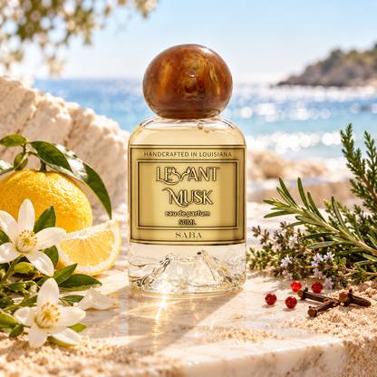A bottle of Saba by Levant Musk Eau De Parfum stands on a stone surface with a beach and ocean in the background, surrounded by citrus, flowers, herbs, and small red berries. The bottle has a round wooden cap and a gold label.