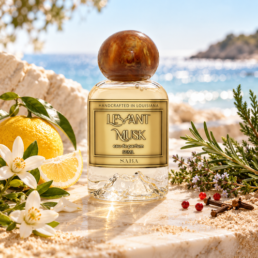 A bottle of Saba by Levant Musk Eau De Parfum stands on a stone surface with a beach and ocean in the background, surrounded by citrus, flowers, herbs, and small red berries. The bottle has a round wooden cap and a gold label.