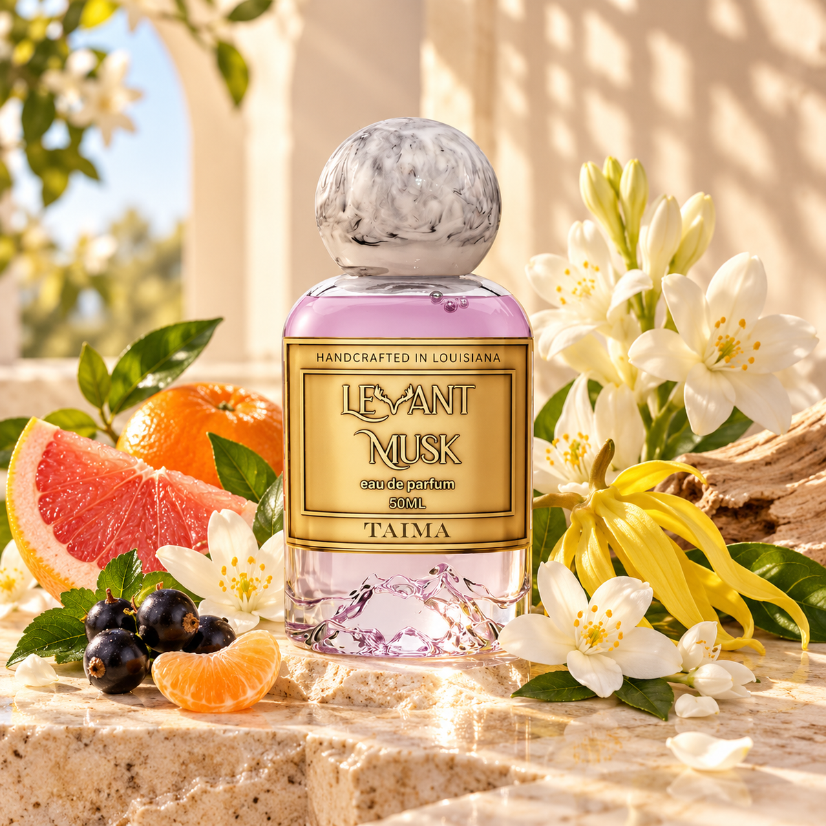 A 50ml eau de perfum bottle labeled TAIMA by Levant Musk, filled with light pink liquid and topped with a marbled gray cap, styled on a stone surface with citrus slices, blueberries, white flowers, and yellow blossoms in a sunlit outdoor setting.